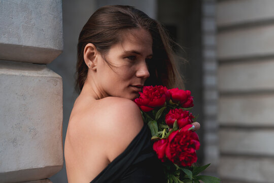 Paeonia flowers held by young woman looking away while leaning against architectural stone column in urban environment