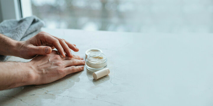 Applying moisturizer to dry hands, next to a jar of cream on a marble surface with a window in the background. The person is focused on moisturizing their hands.