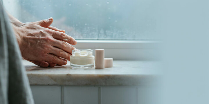 Hands applying cream to the skin, with a window in the background
