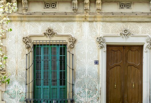Intricate sgraffito patterns adorn a historic building facade in Sitges, Spain, highlighting the detailed artistry of Catalan Modernism around a window with green shutters and a wooden door