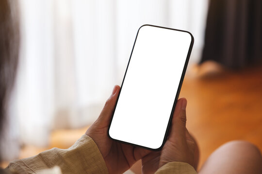 Mockup image of a woman holding and using mobile phone with blank desktop white screen at home