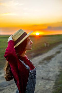 Young Woman in Straw Hat at Sunset. Young woman wearing a straw hat standing on a rural road during a warm countryside sunset.