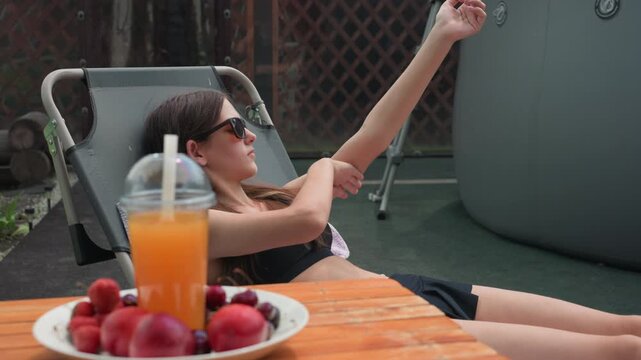Young woman relaxes in backyard applying sunscreen to her arm while seated on outdoor chair. plate of fresh fruit and drink are placed nearby with background featuring garden elements
