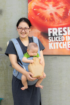 Mother and baby shopping lifestyle concept, happy Asian mother with glasses carrying infant in baby carrier holding food paper bag, representing urban parenting, family errand, and takeaway.