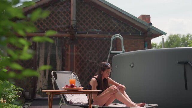 Young woman relaxes outdoors near hot tub adjusting her posture on lounge chair. wooden table beside her holds fruit and orange drink in plastic cup with straw. background features greenery