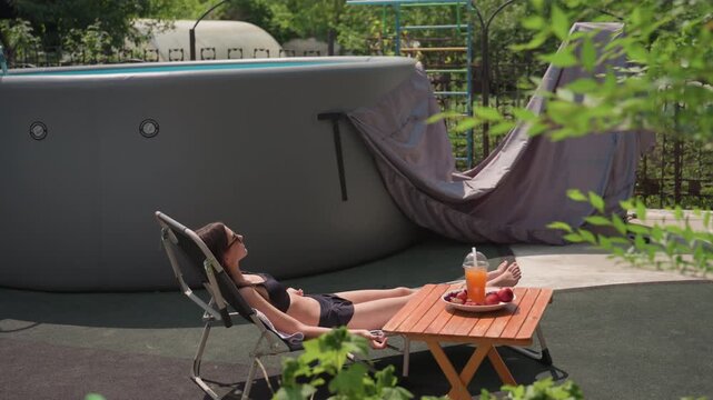 Woman in black swimsuit relaxes on reclining chair outdoors near above ground pool. wooden table beside her holds fruit and orange juice with green foliage partially framing scene. setting