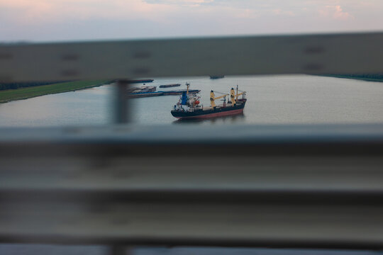 Cargo vessel sailing along a wide river, viewed through bridge railings on a cloudy day, showcasing transport, trade, and industry.