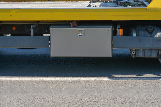A side view close up shot of a gray stainless steel tool storage box mounted under the yellow chassis of a vehicle on an asphalt road.