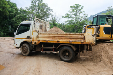 Old dump truck carrying sand beside construction excavator on muddy outdoor worksite, with dense forest and mountain scenery creating active industrial landscape © CoreRock