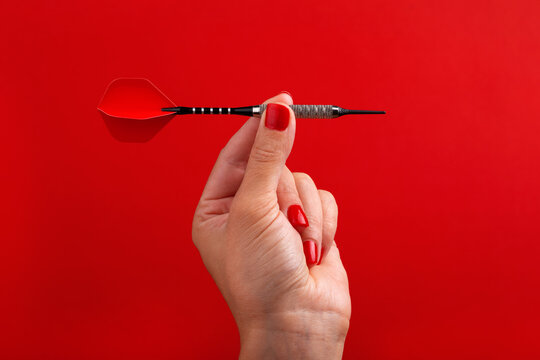 Close-up of a woman's hand with red nails holding a dart against a vibrant red background. Concept of precision, focus, and target achievement.