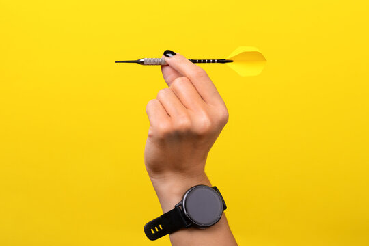 Close-up of a hand with smartwatch holding a dart against a solid yellow background, symbolizing focus and precision.