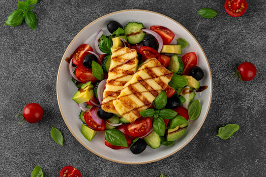 Grilled halloumi salad with tomatoes, cucumber, avocado, olives, basil and balsamic glaze on a plate on a concrete background. Healthy Mediterranean food. Top view, copy space.