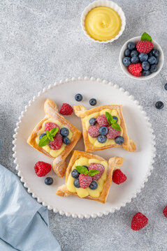 Danish puff pastry cakes with custard cream and fresh berries, sprinkled with powdered sugar on a white plate on a concrete background.