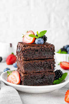 A stack of chocolate brownies with chocolate glaze, fresh berries and mint leaves on top on a white plate on a gray concrete background. Delicious dessert.