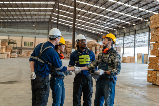 Lumber surrounds factory workers showing teamwork during a team meeting as two men shake hands beside two smiling colleagues.