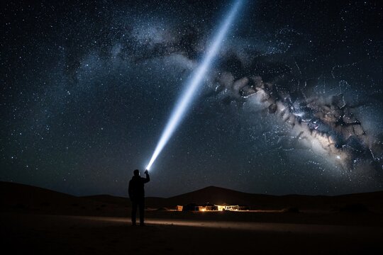 Man holds bright flashlight towards Milky Way galaxy in Moroccan desert at night. Silhouette person shines light beam into starry sky above sand dunes. Small desert camp glows in distance under vast