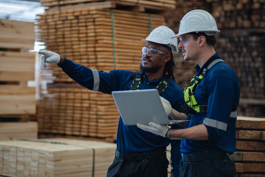 Checking lumber inventory in the warehouse, two men hold a laptop while one points away beside tall stacks of timber.