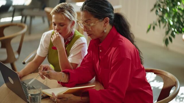 Seated women share business ideas over a laptop in a coworking cafe, showing collaboration and teamwork as they work together with notes and planning in a bright indoor setting.