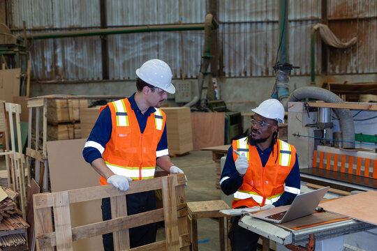 During woodworking, a carpenter discusses plywood with his colleague in a wood workshop beside a laptop, holding a pencil.