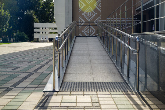 Accessible building entrance ramp made of stone tile with shiny stainless steel handrails for wheelchair users.