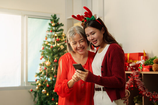 Asian mother and adult daughter using smartphone near Christmas tree for holiday connection