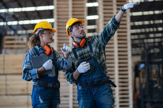 Two factory workers show teamwork at a team meeting, pointing upward together with a tablet beside stacked wooden pallets.