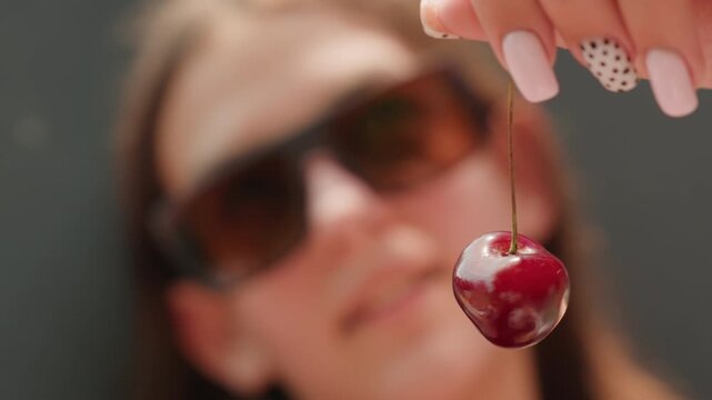Woman holds single red cherry close up showcasing its glossy texture and stem. Her hand with manicured nails featuring one dot pattern is visible as woman presents fruit against blurred