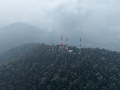 Aerial view of telecommunication towers standing on a lush forested hilltop under a cloudy sky in Bukit Mertajam, Penang, Malaysia.