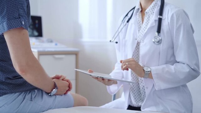 Doctor woman is taking notes on a tablet computer while a male patient describes his symptoms in clinic procedure cabinet. Medicine service concept