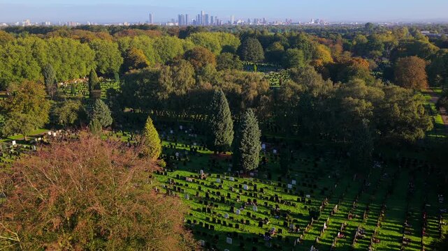 Sunlit aerial video looking over tree-lined avenues and headstone sections of Manchester Southern Cemetery toward the Manchester city centre skyline.