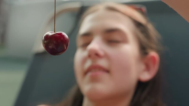 Young woman observes single red cherry hanging on string in close up view. background remains softly blurred emphasizing shiny surface and rich color of fruit as it gently sways. Soft