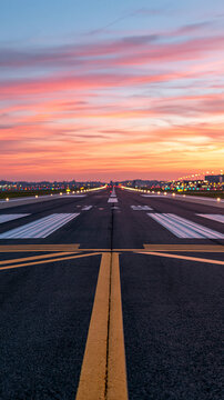 Empty runway at an airport in the evening with amber light grazing the asphalt centerline markings receding beneath a rose colored sunset sky distant edge lights beginning