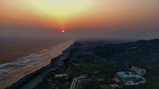 Aerial view of Parangtritis Beach at sunset with golden light reflecting on the waves, dark sand coastline, and surrounding lush hills in Kecamatan Kretek, Daerah Istimewa Yogya, Indonesia.