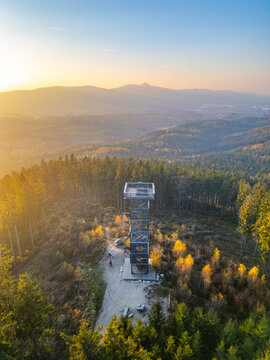 Aerial view of Cisaruv kamen lookout tower surrounded by autumn forest and hills during golden hour in Liberec 30, Liberec Region, Czechia.