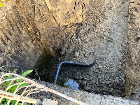 Excavation site with deep trench dug in dark earth, showing exposed soil layers, water and corrugated plastic drainage pipe