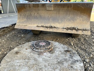 Bulldozer blade of heavy machinery closeup over dirt and gravel next to sewer manhole cover on construction site © grooveisintheheart