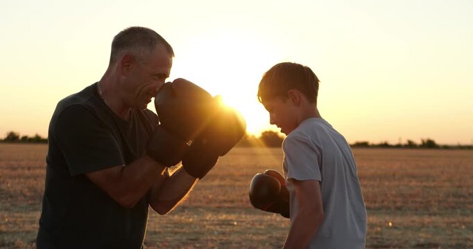 Father and son practice boxing together in field at sunset. They train with focus, teamwork, and affection. Golden light creates family fitness scene for sports, parenting, and healthy living.