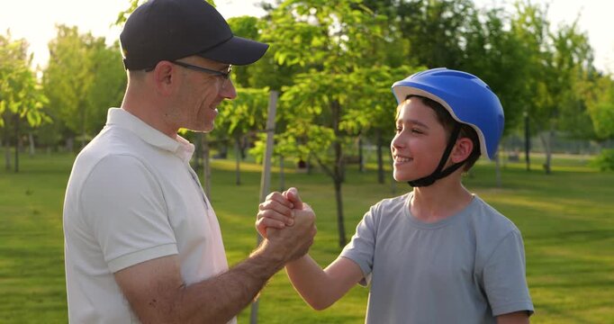 Father and son share moment in sunny park. Boy wears helmet and holds scooter while father offers encouragement. Happy family bond promotes outdoor activity, safety, and active parenting.