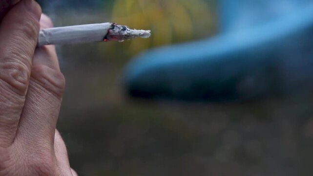 Man holding a lit cigarette with smoke rising in an outdoor setting