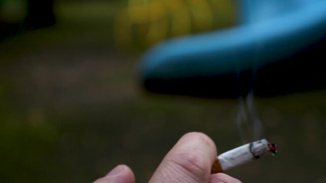 Man holding a lit cigarette in his hand with smoke rising from the burning tip outdoors
