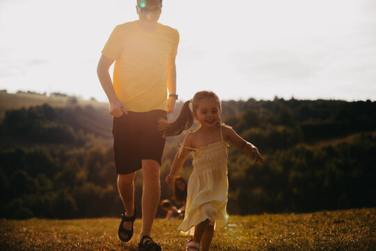 Father And Daughter Running Through Sunlit Field At Sunset In Warm Summer Playful Moment