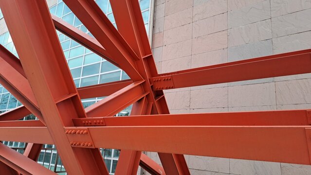 close-up of red steel beams converging at bolted joint connection with stone tile wall and glass building in background