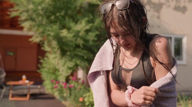 Young woman with wet hair dries herself outdoors wearing black sports bra and holding light purple towel. scene features green foliage pink flowers outdoor table with items soft natural