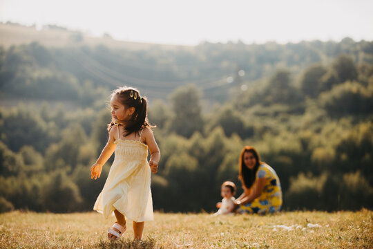 Little Girl Playing in Sunlit Meadow While Mother Watches With Baby on Summer Countryside Afternoon
