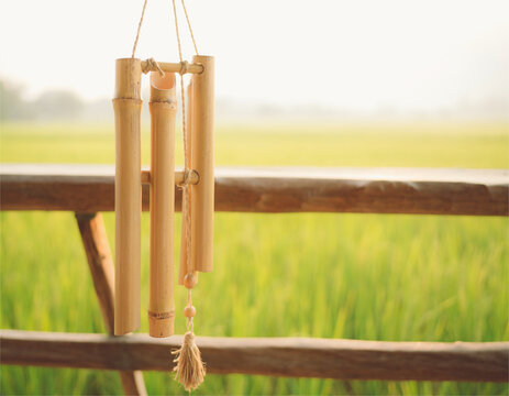 Bamboo wind chime hanging on a rustic wooden railing overlooking a blurred green rice field in soft morning light with copy space