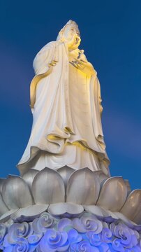 A tall white statue of a goddess stands on a lotus pedestal against a blue sky. The statue is illuminated, showcasing intricate details and serene expression.