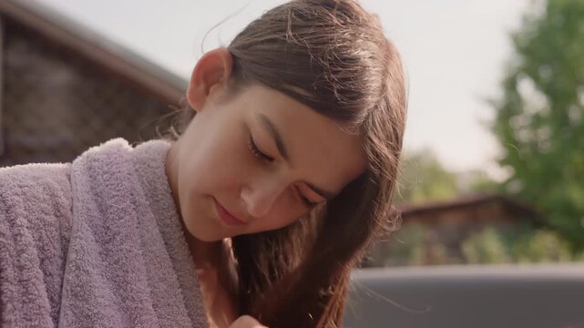 Young woman wearing purple robe combs another woman hair with pink comb and scrunchie focusing on gentle interaction between them. scene is set outdoors in soft daylight featuring natural