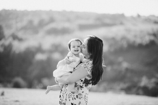 Mother Kissing Laughing Baby While Holding Her Outdoors In Black And White Portrait