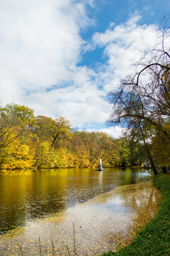 Bright autumn landscape with golden trees surrounding a peaceful lake, tranquil water, elegant fountain, cozy shoreline, colorful fall foliage, blue sky and clouds creating a serene retreat
