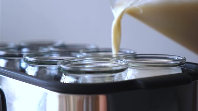 Cream pours into glass jars on tray from spout of kitchen machine. Filling process continues smoothly as jars wait beneath stream, suggesting small batch food production with careful preparation.